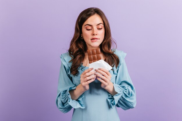 Portrait of funny brunette woman biting her lip in anticipation of tasty lunch. Girl in blue dress looks at delicious chocolate.