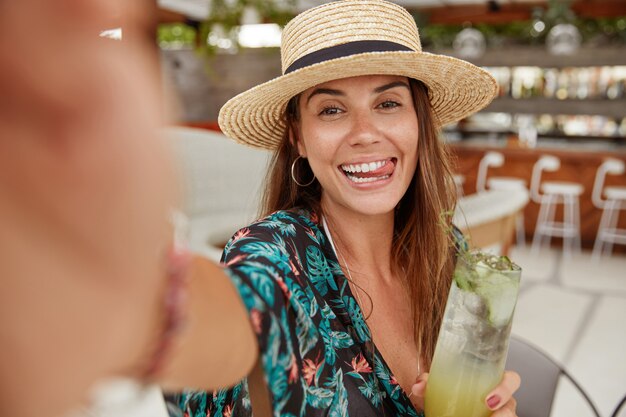 Portrait of funny brunette woman wears straw hat, poses for selfie, shows tongue, drinks refreshing cocktail, stands against cafe interior. Beautiful young female model enjoys summer holiday