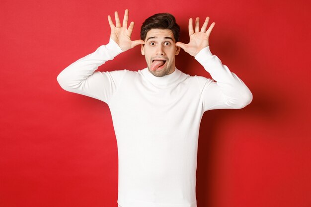 Portrait of funny caucasian guy showing tongue and making faces wearing white sweater standing again...