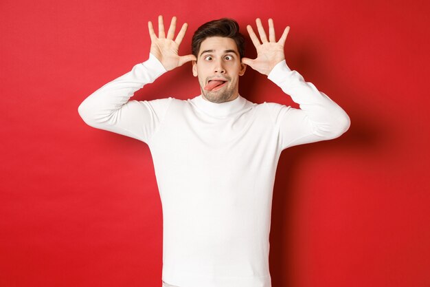 Portrait of funny caucasian guy showing tongue and making faces wearing white sweater standing again...