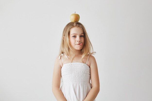 Portrait of funny cheerful girl with blond hair in white dress holding apple on head, showing tongue, making silly expressions.