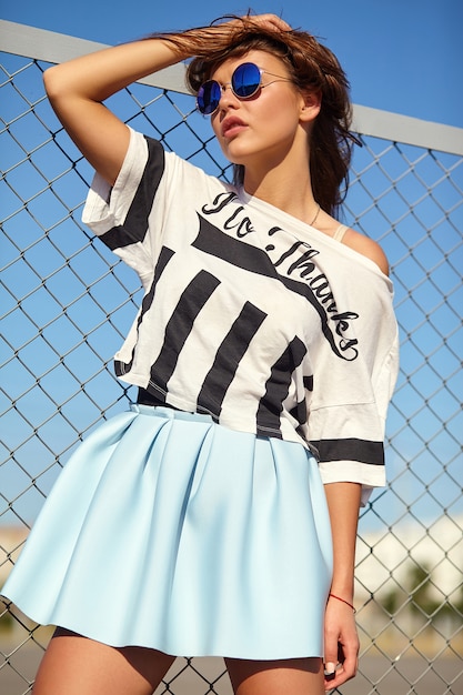 Free photo portrait of funny crazy glamor stylish smiling beautiful young woman model in bright hipster summer casual clothes posing in the street behind iron grating and blue sky