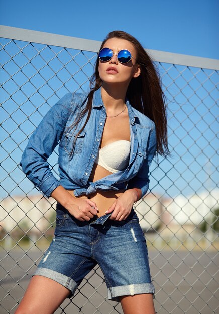 Portrait of funny crazy glamor stylish smiling beautiful young woman model in bright hipster summer casual jeans clothes posing in the street behind iron grating and blue sky