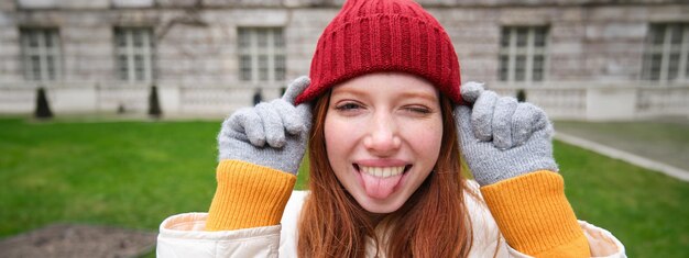 Portrait of funny and cute redhead girl puts on red hat shows tongue and winks at camera smiles