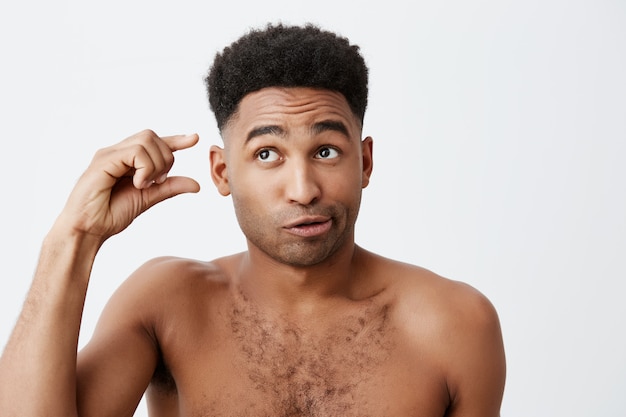 Portrait of funny dark-skinned american guy with curly hair and without clothes looking aside with silly and cynic expression, gesticulating with hand. People's emotions.