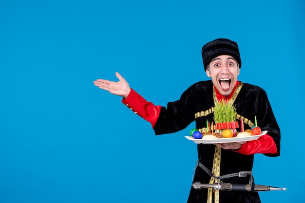 Portrait of funny emotional young adult in traditional dress showing tray filled with national confectionery pointing something on blue wave background