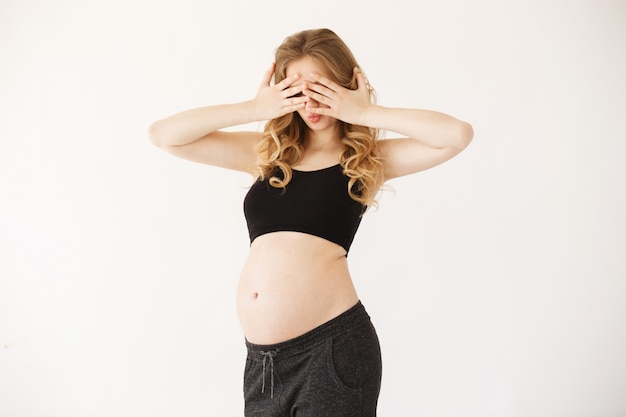 Free photo portrait of funny european mother with blond curly hair in black sport suit with opened pregnant belly, clothing eyes with hands, having surprised expression