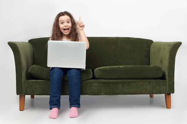 Portrait of funny excited little girl in jeans sitting on sofa with portable computer on lap, exclaiming excitedly and raising finger.