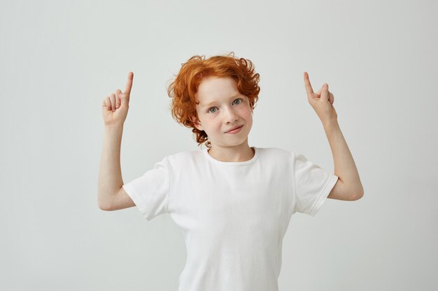 Portrait of funny ginger child with freckles pointing upside on white wall with relaxed expression