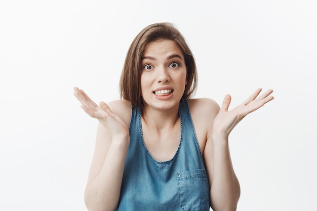 Portrait of funny good-looking european student girl with dark long hair in blue casual clothes  with confused expression, gesticulating with hands showing she can't answer question f