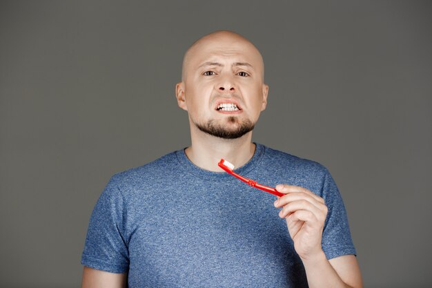 Portrait of funny handsome man in grey shirt brushing teeth over dark wall