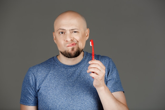 Portrait of funny handsome man in grey shirt holding toothbrush over dark wall