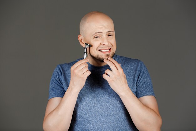 Portrait of funny handsome man in grey shirt shaving over dark wall