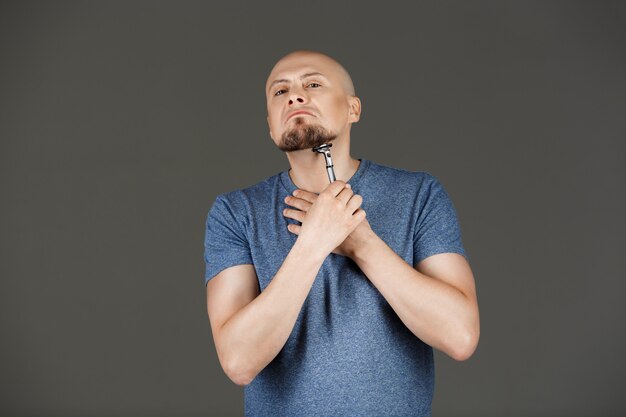 Portrait of funny handsome man in grey shirt shaving over dark wall