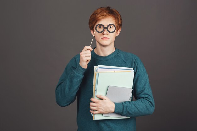 Portrait of funny handsome redhead teen in green fashionable sweater holding notebooks in hand, looking with popped up eyes through paper glasses on stick.