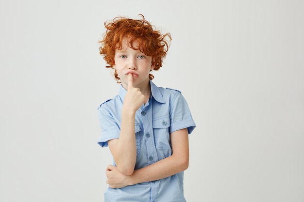 Ritratto di bambino divertente con capelli ondulati allo zenzero e lentiggini tenendo il dito in bocca con l'espressione del viso annoiato