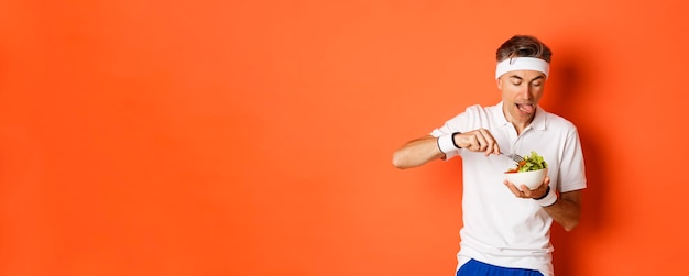 Ritratto di divertente sportivo di mezza età in uniforme da allenamento che lecca le labbra mangiando una deliziosa insalata dopo