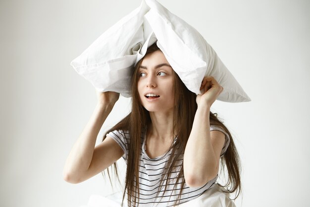 Free photo portrait of funny playful beautiful young 20 year old woman with long loose dark hair wearing striped t-shirt indoors, looking sideways with mysterious smile, holding white pillow over her head