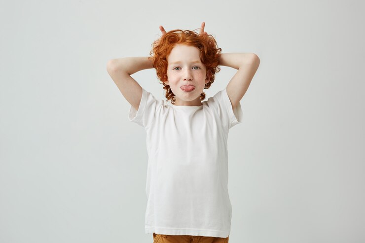 Free Photo portrait of funny redhead little boy with freckles having fun indoors, sticking out her tongue and making horns with fingers.