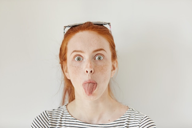 Portrait of funny redhead young Caucasian woman with freckles having fun indoors, sticking out her tongue. Close up of teenage girl wearing trendy sailor shirt making faces at white wall