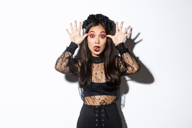 Portrait of funny and silly asian woman in witch costume celebrating halloween, mocking someone, showing faces and sticking tongue, standing over white background.