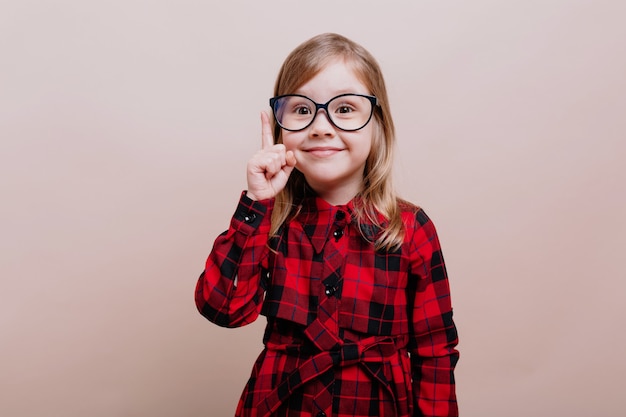 재미 있은 똑똑한 어린 소녀의 초상화는 안경을 착용하고 체크 무늬 셔츠는 한 손가락을 들고 정면에 미소 짓습니다.