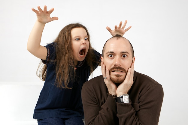Foto gratuita ritratto di divertente giovane maschio barbuto tenendo le mani sul viso, terrorizzato dalla sua piccola figlia che in piedi dietro di lui, gridando