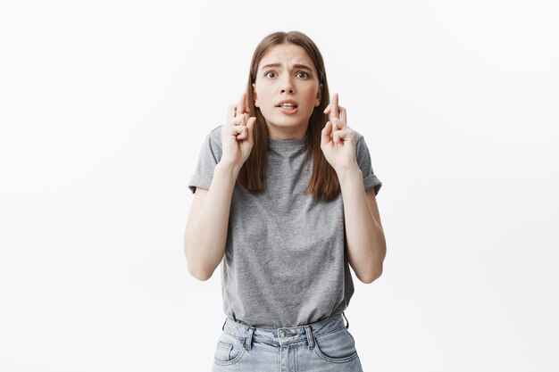 Portrait of funny young beautiful caucasian student girl with dark hair in casual clothes crossing fingers,  with pleasing face expression asking for luck on test in university