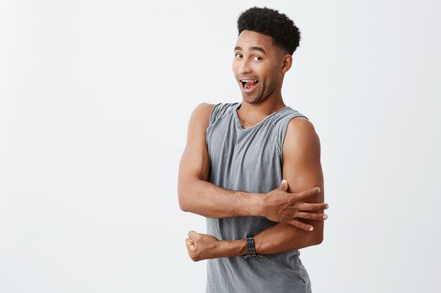 Portrait of funny young dark-skinned african male with curly hair in grey sporty wear touching his muscles with hand, looking in camera with silly expression. Sport concept
