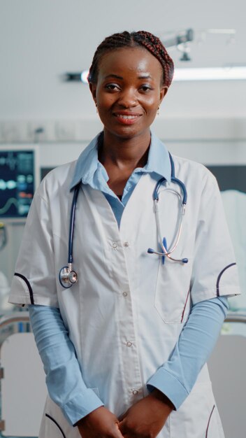 Portrait of general practitioner standing in hospital ward, ready to treat patient with disease. Woman doctor with stethoscope and white coat working with medical equipment at clinic.