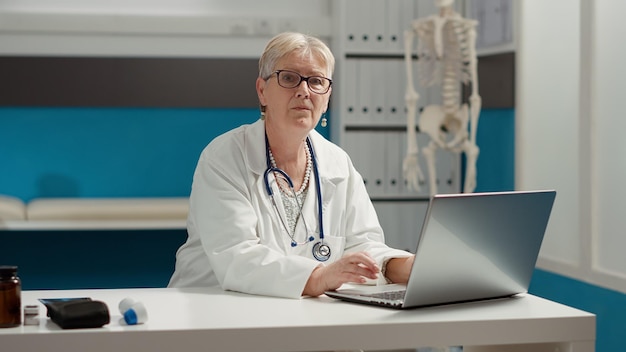 Portrait of general practitioner working on laptop in medical office, preparing to attend appointment. Doctor with health care expertise using hospital system to plan checkup consultation.