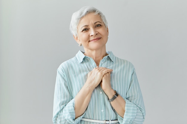 portrait of generous kind mature senior female in stylish shirt holding hands clasped on her chest, feeling grateful for great present on her birthday. elderly woman expressing appreciation