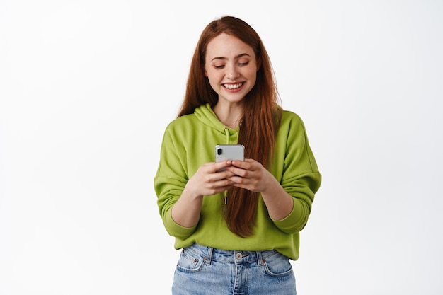 Girl turn attention at camera, distracted from smartphone smiling friendly  and happily holding mobile phone as, taking selfie in mirror, showing  friends new outfit, standing in dressing room of mall. Photos