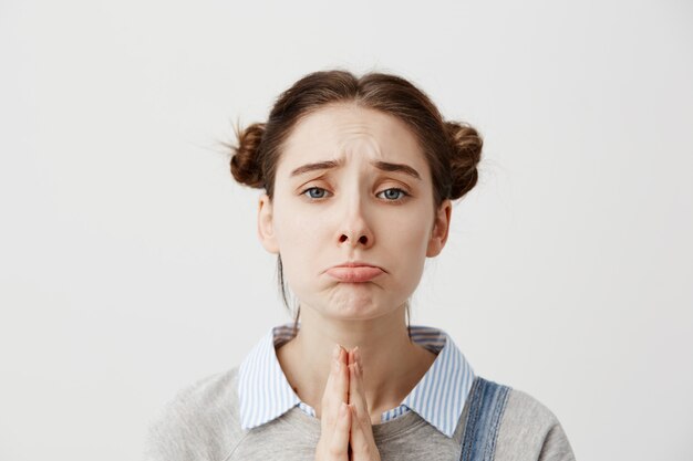 Portrait of girl 20s with upset look begging for mercy being disappointed with praying gestures. Capricious female with brown hair in double buns pouting having hopeful expression. Close up