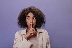 Free photo portrait of girl in beige jacket puts finger to her mouth young girl in stylish outfit keeps secret and posing on purple background