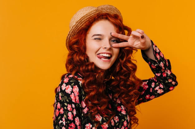 Free photo portrait of girl fooling around on orange space. lady in floral blouse winks and shows peace sign.