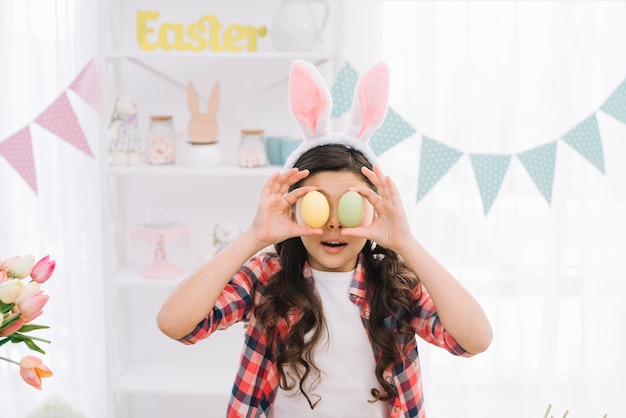 Free photo portrait of a girl holding easter eggs in front of her eyes on easter day at home