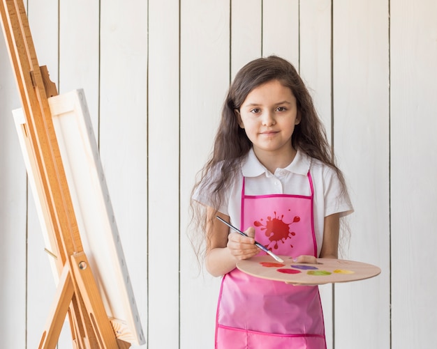 Free photo portrait of a girl holding paint brush and palette in hand near the easel