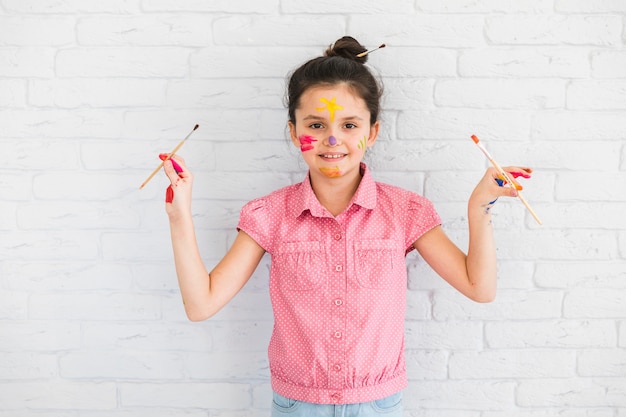 Portrait of a girl holding paint brushes in hand standing in front of white brick wall