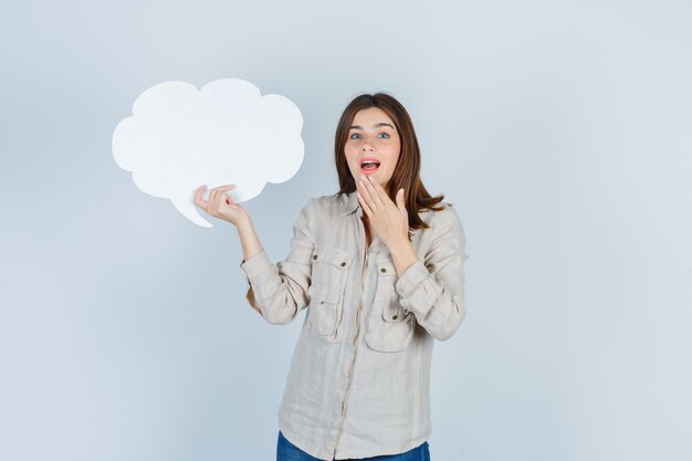 Portrait of girl holding speech bubble, keeping hand on chin in shirt and looking surprised