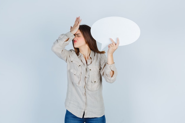 Free photo portrait of girl holding speech bubble, keeping hand on forehead in shirt and looking forgetful