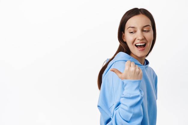 Portrait of girl in hoodie, pointing left and looking at sale promo text, showing banner, smiling and laughing happy, standing against white background