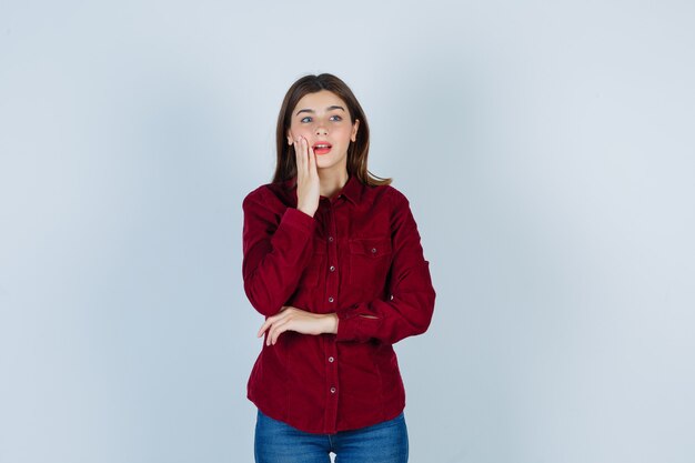 Portrait of girl keeping hand on cheek in burgundy blouse and looking curious