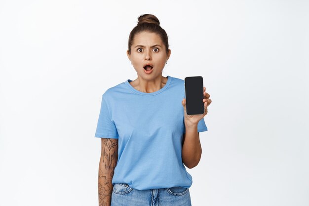 Portrait of girl looks shocked and shows mobile phone screen, standing over white background