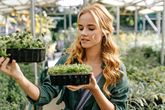 緑の小さな植物と2つのプラスチック製のポットを持って外の女の子の肖像画。若い女性の植物学者は緑を研究します。