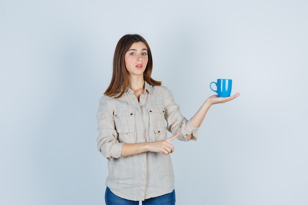 Foto gratuita ritratto di ragazza che indica una tazza di tè in camicia e sembra esitante