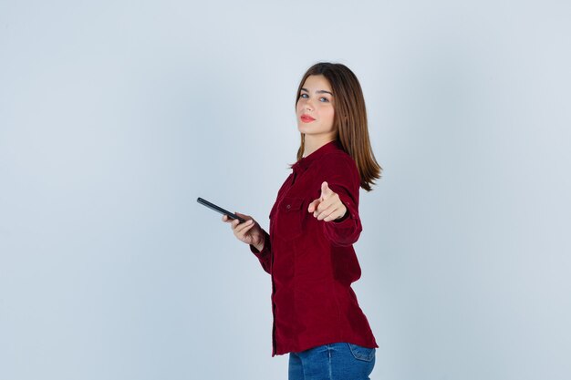 Портрет девушки, указывающей вперед, держащей смартфон в бордовой рубашке и уверенной в себе