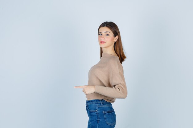 Портрет девушки, указывающей куда-то, стоя боком в свитере, джинсах и весело выглядящей