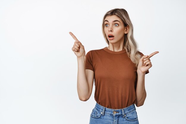 Portrait of girl points sideways at two options looking at left side with surprised impressed face expression standing against white background