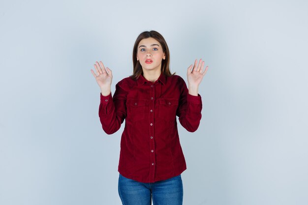 Portrait of girl raising hands in surrender gesture in burgundy blouse and looking scared